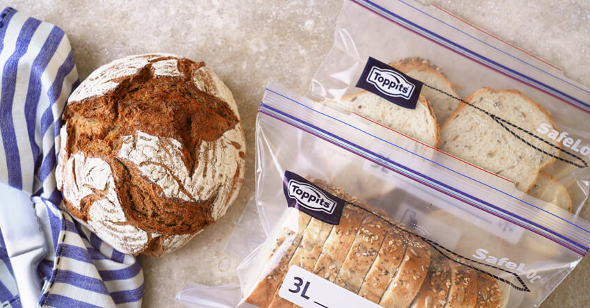 Ein Laib Brot und aufgeschnittenes Brot in zwei Toppits SafeLoc Gefrierbeuteln liegen auf einer hellen Arbeitsfläche neben einem blau-weiß gestreiften Tuch